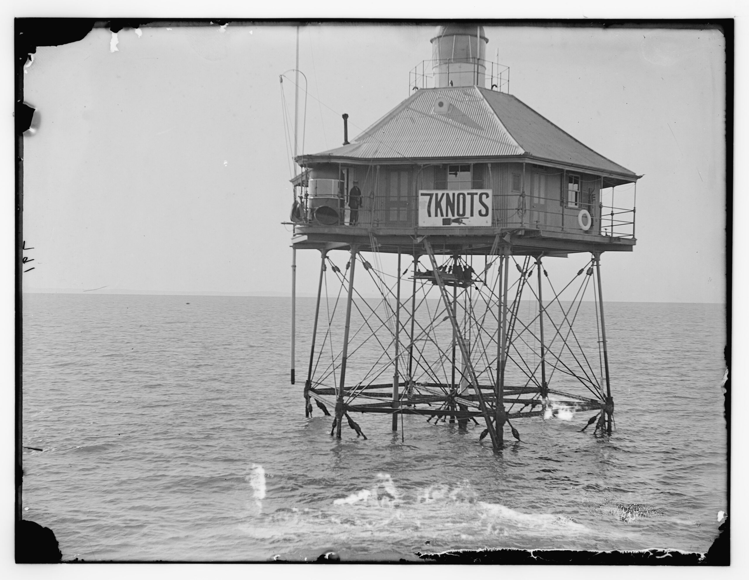 Black and white photograph of a screw-pile lighthouse standing in open water, a painted 7 KNOTS sign on its side and a keeper on the gallery.